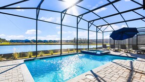 Piscina com vista para um lago e edifícios ao longe, rodeada por uma área coberta com espreguiçadeiras e um guarda-sol.