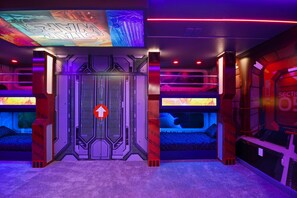 Quarto com design futurista, destacando uma porta central com sinal de seta indicativo, paredes com padrões hexagonais e uma tela grande exibindo imagem com tema espacial.
