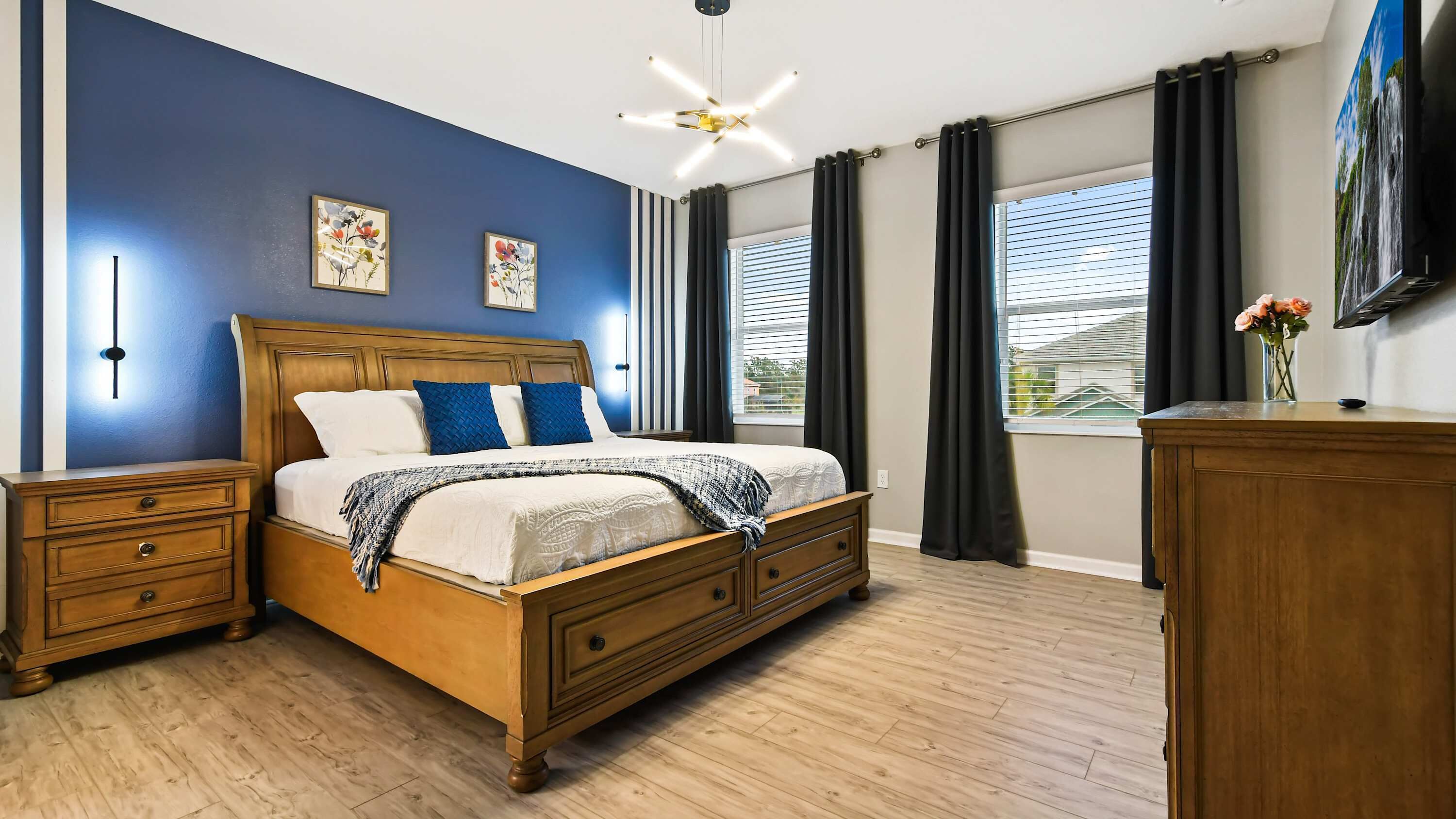 Une chambre à coucher avec un lit en bois, deux tables de chevet, une commode, un ventilateur de plafond, une fenêtre avec des rideaux et un téléviseur fixé au mur.