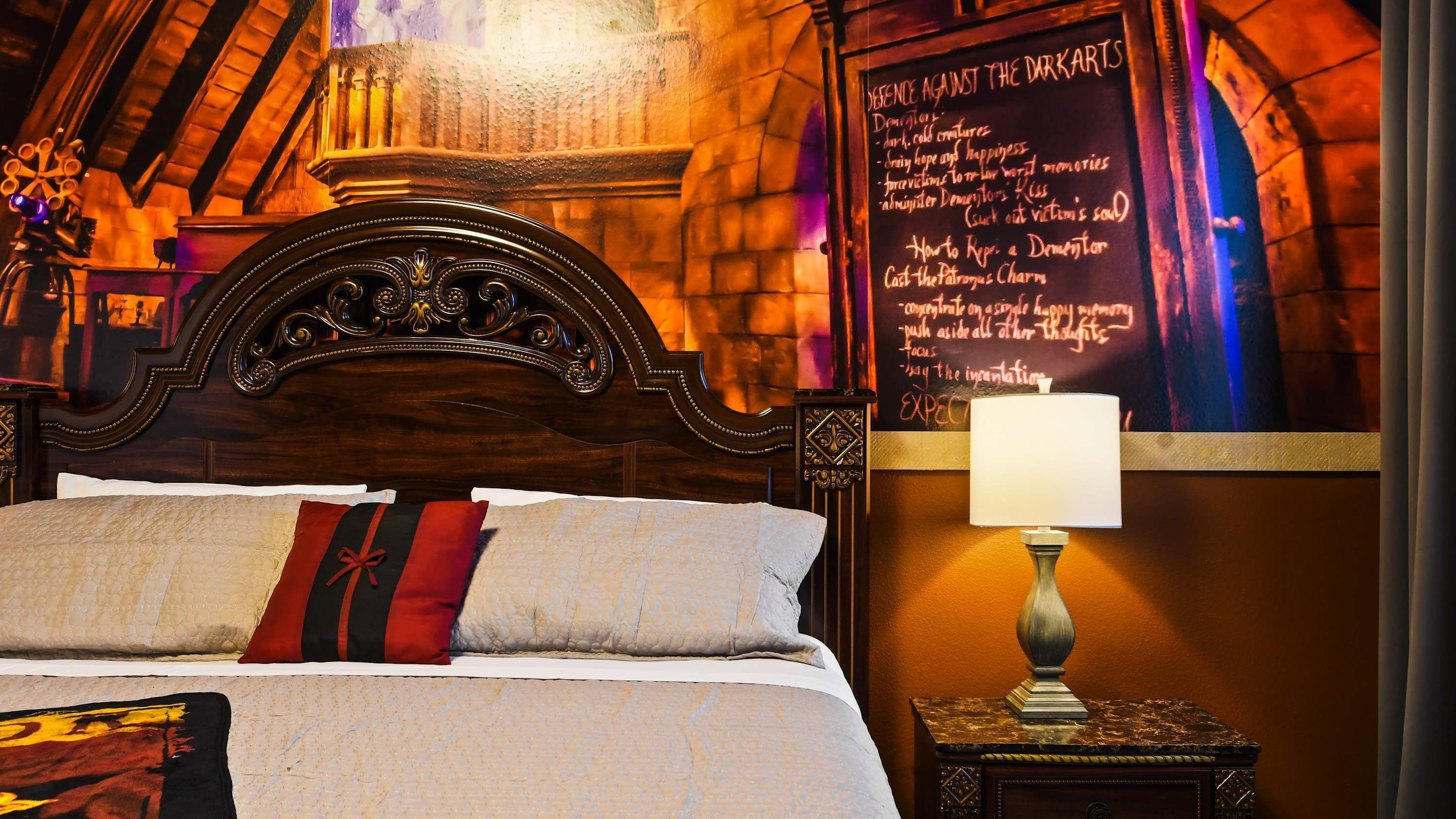 A hotel room with a wooden headboard, a bed with a white and red pillow, a nightstand with a lamp, and a wall with a chalkboard listing 'Defence Against the Dark Arts' topics.