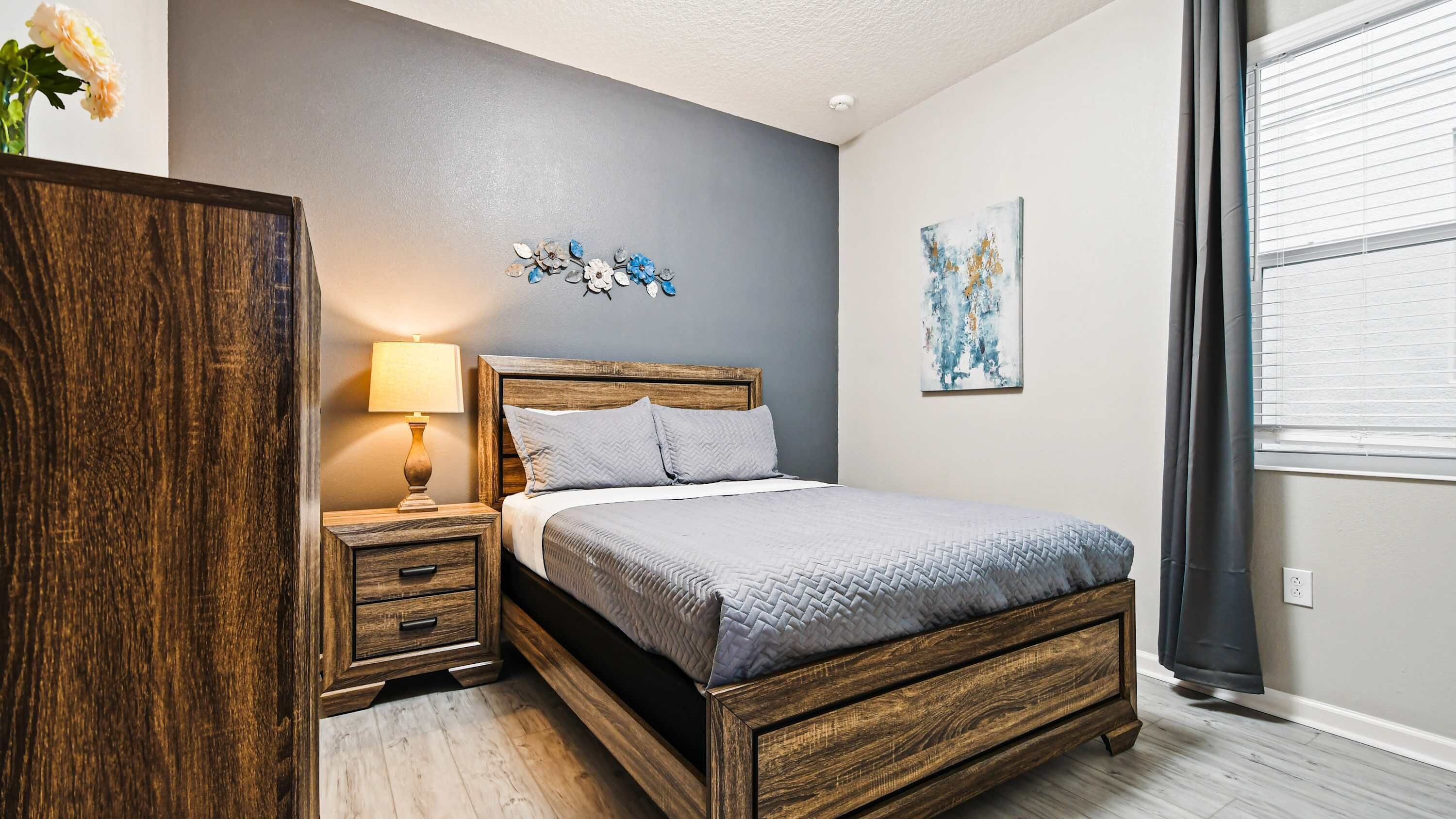 A bedroom with a wooden bed, a nightstand with a lamp, a dresser, and a window with blinds.