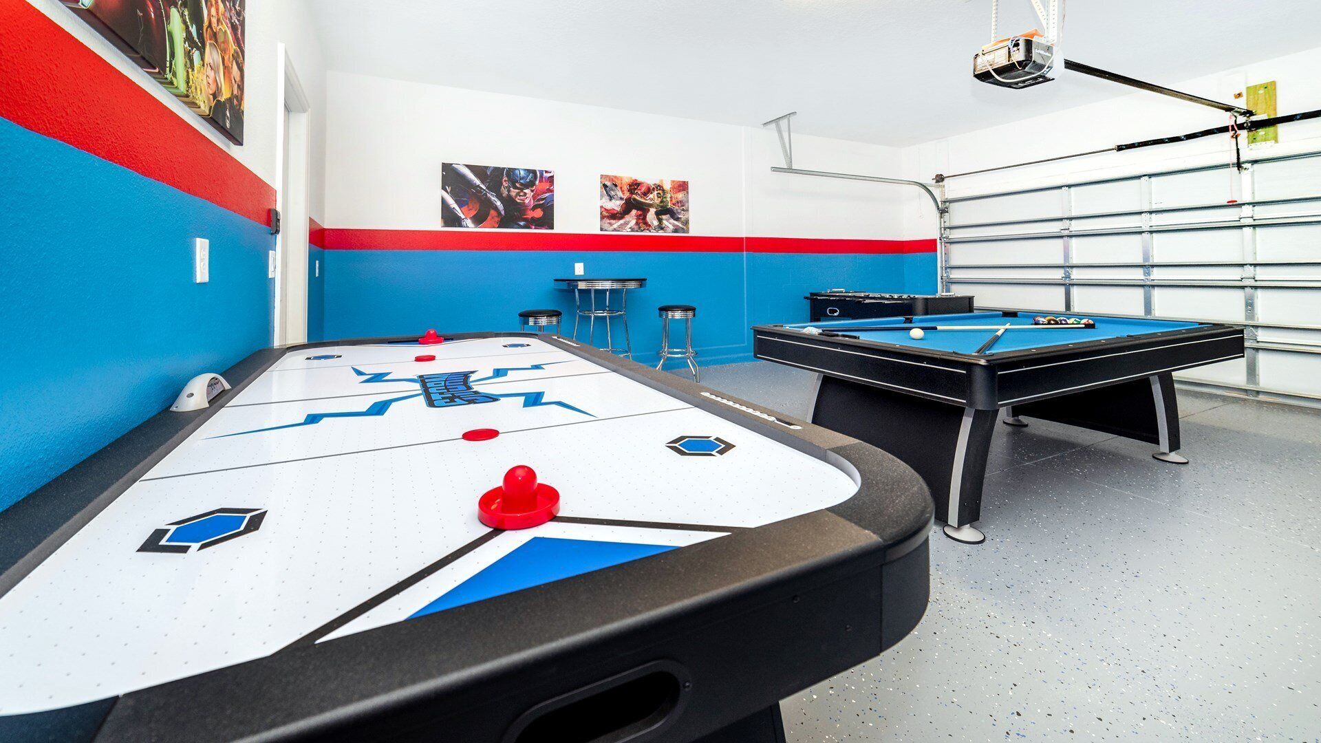 A pool table with a blue and white design, a second pool table, and a foosball table in a room with blue and white walls and a garage door in the background.