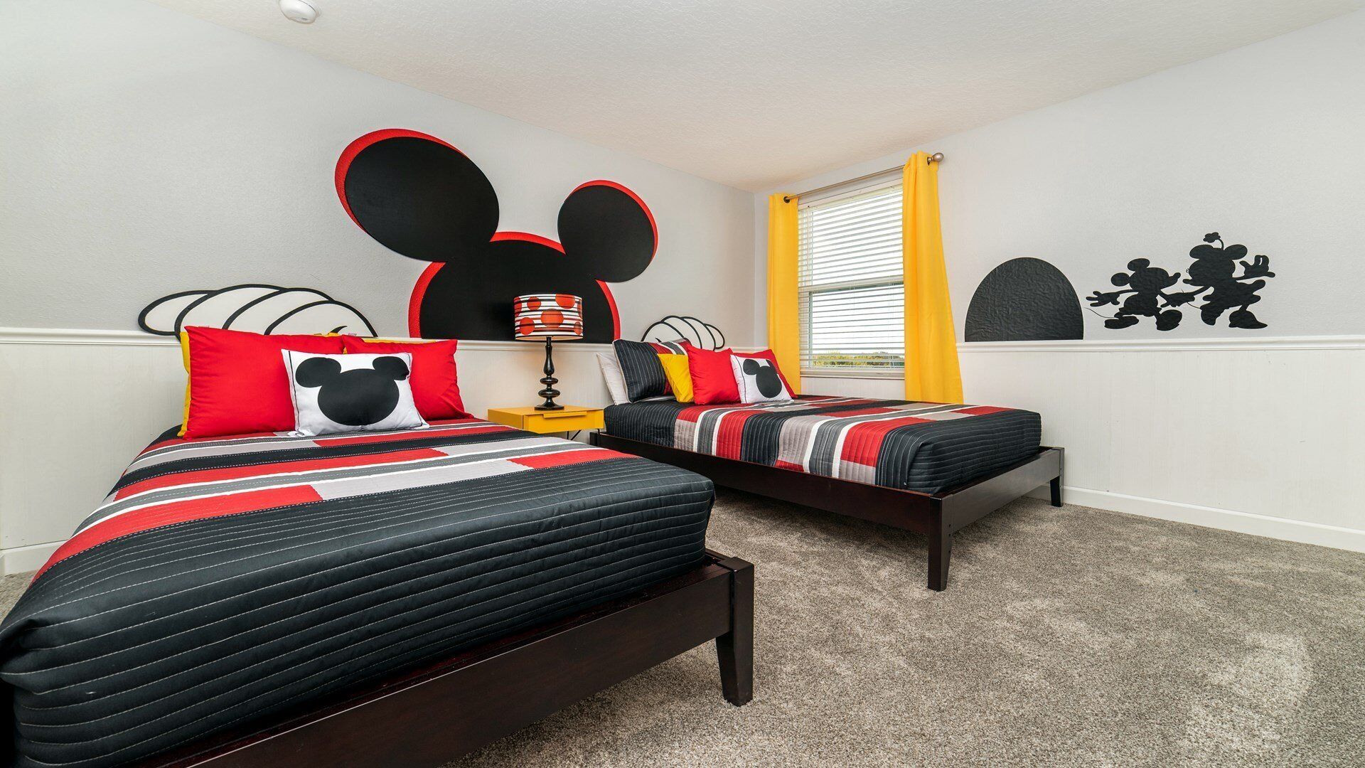 A bedroom with two beds, each with a Mickey Mouse themed headboard, a nightstand with a lamp, and a window with yellow curtains.