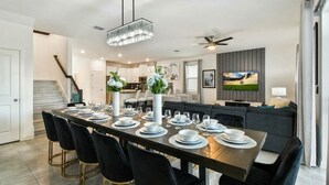 Un espace de repas moderne avec une longue table dressée pour six personnes, un escalier, une cuisine et un coin salon avec un canapé et un téléviseur.