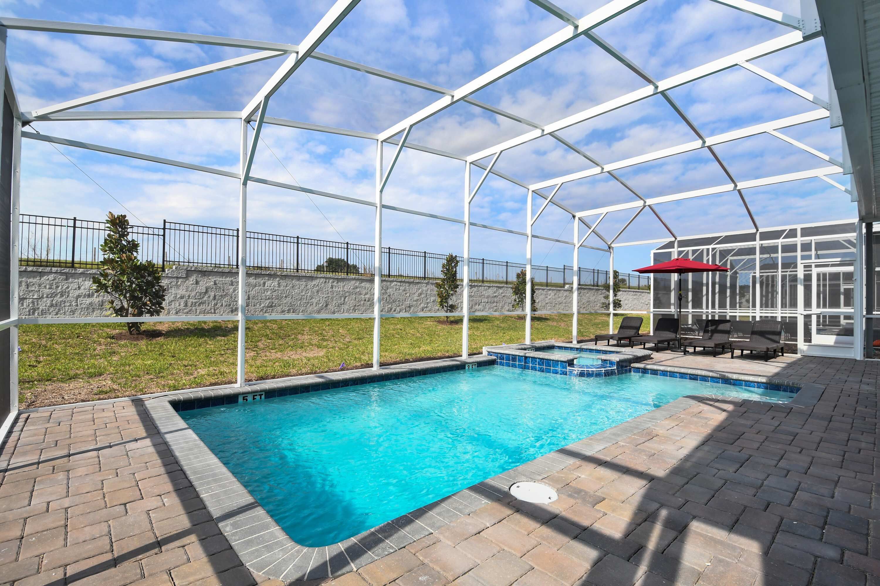 A covered outdoor swimming pool with a brick patio, lounge chairs, and a red umbrella.