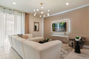 Un salon moderne avec un canapé d’angle, un téléviseur à écran plat fixé au mur, un miroir décoratif et un lustre.