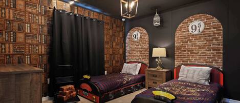 Una habitación con pared de ladrillo, dos camas con ropa de cama temática, una estantería y un carrito de equipaje.