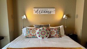 Cama bem arrumada com edredom branco e manta amarela texturizada. Há dois travesseiros decorativos com padrões elaborados e um letreiro acima da cama com a mensagem 'sonhos doces'.