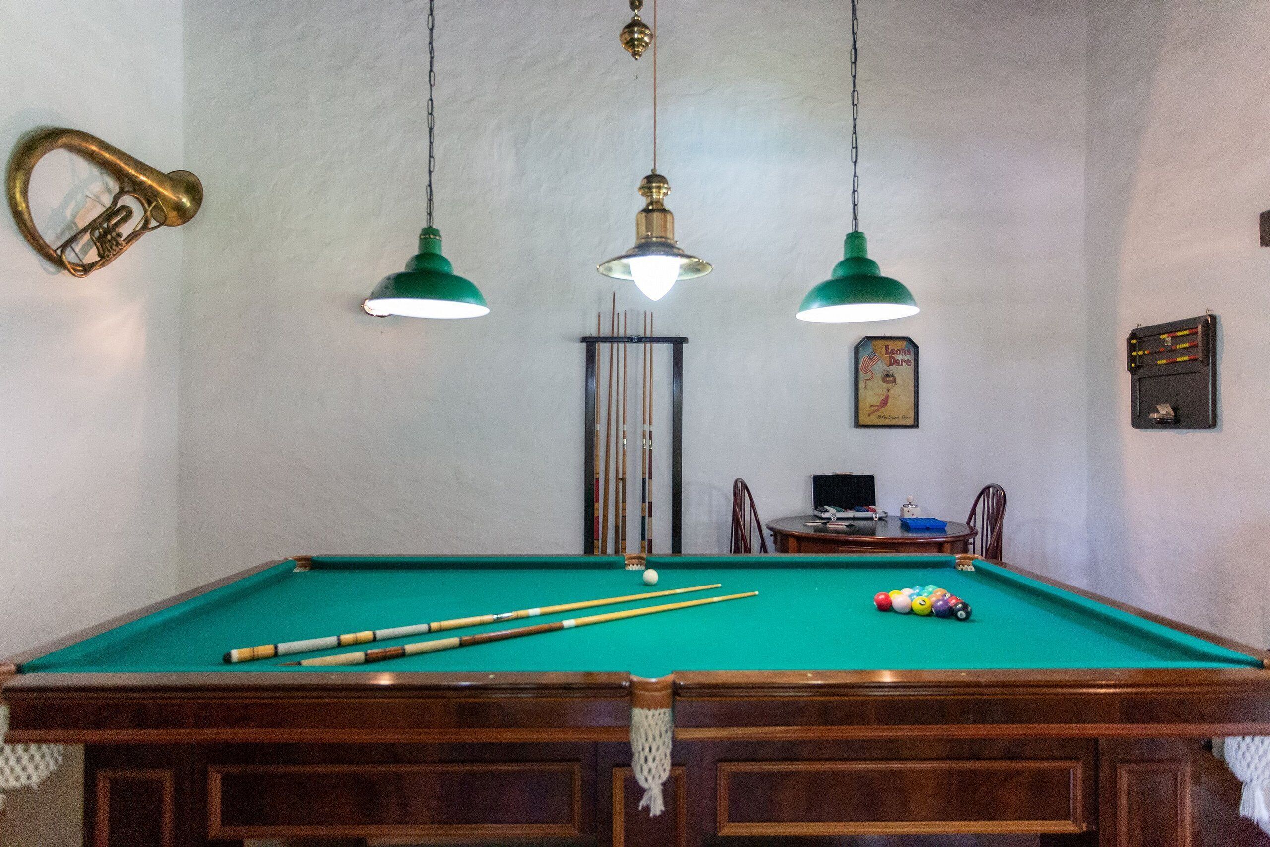 Une table de billard recouverte de feutre vert, deux lampes suspendues vertes, un loquet de porte en laiton et un tableau encadré accroché au mur.