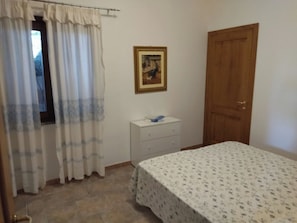 Un dormitori amb un llit, una tauleta de nit, una porta de fusta i una finestra amb cortines.
