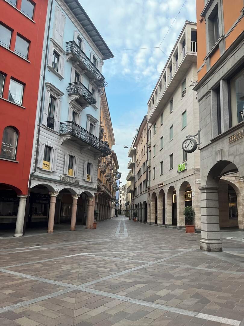 Una calle peatonal con edificios históricos, incluyendo un edificio rojo con un letrero 'TOURBILLON' y un reloj en un edificio con un letrero 'PAX'.