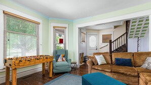 Un salon chaleureux avec un baby-foot, un fauteuil vert, un canapé marron agrémenté de coussins bleus et à motifs, et un pouf bleu.