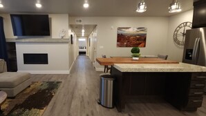 Ein modernes Wohnzimmer mit Sofa, Couchtisch und einem an der Wand befestigten Fernseher. Es gibt einen Essbereich mit einem Holztisch und Stühlen, sowie eine Küchenzeile mit Kühlschrank und einer Wanduhr.