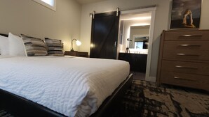 Ein ordentlich bezogenes Bett mit weißer Bettwäsche, ein Holzkasten mit Schubladen, eine Schreibtischlampe und ein Spiegel an der Wand.