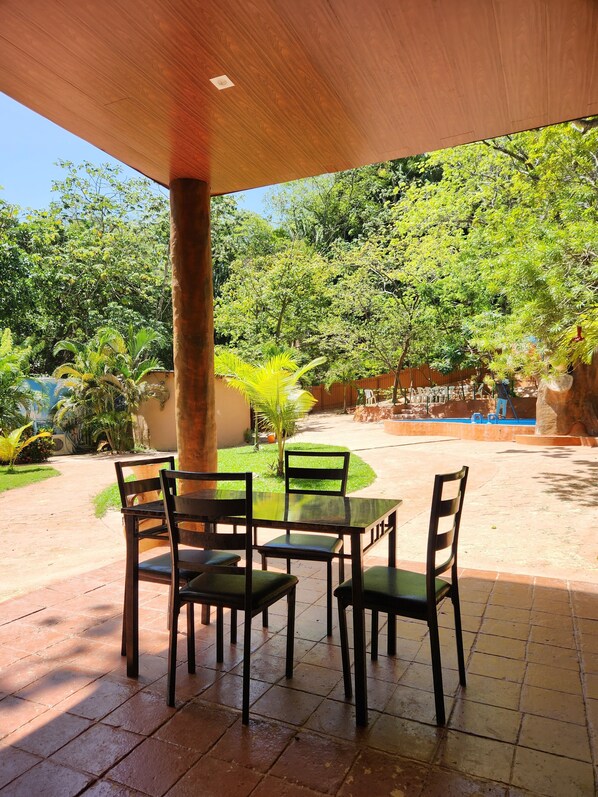An outdoor patio with a wooden table and chairs, surrounded by lush greenery and a clear blue sky.