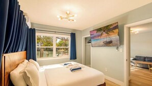 Une chambre à coucher avec un lit, une fenêtre avec des rideaux, un tableau représentant une scène de plage et un canapé dans la pièce adjacente.