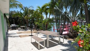 Une terrasse extérieure avec une table et des bancs, entourée de palmiers et offrant une vue sur une étendue d’eau.