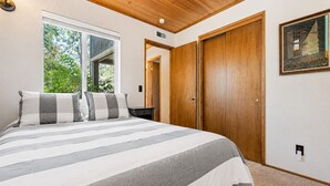 A bedroom with a large bed, a wooden wardrobe, a nightstand, and a window with a view of greenery. - Cub Run Cabin (Big Bear Lake)