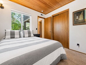 A bedroom with a large bed, a wooden wardrobe, a nightstand, and a window with a view of greenery. - Cub Run Cabin (Big Bear Lake)