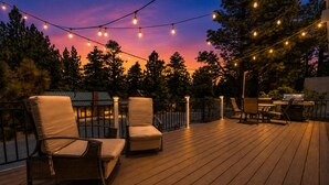 An outdoor deck with wooden flooring, two cushioned chairs, a grill, and string lights against a twilight sky. - Cub Run Cabin (Big Bear Lake)