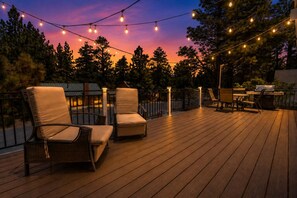 An outdoor deck with wooden flooring, two cushioned chairs, a grill, and string lights against a twilight sky. - Cub Run Cabin (Big Bear Lake)