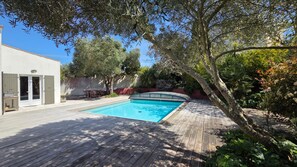 Una piscina rectangular con cubierta de madera, rodeada de árboles y un edificio blanco con puerta de vidrio.