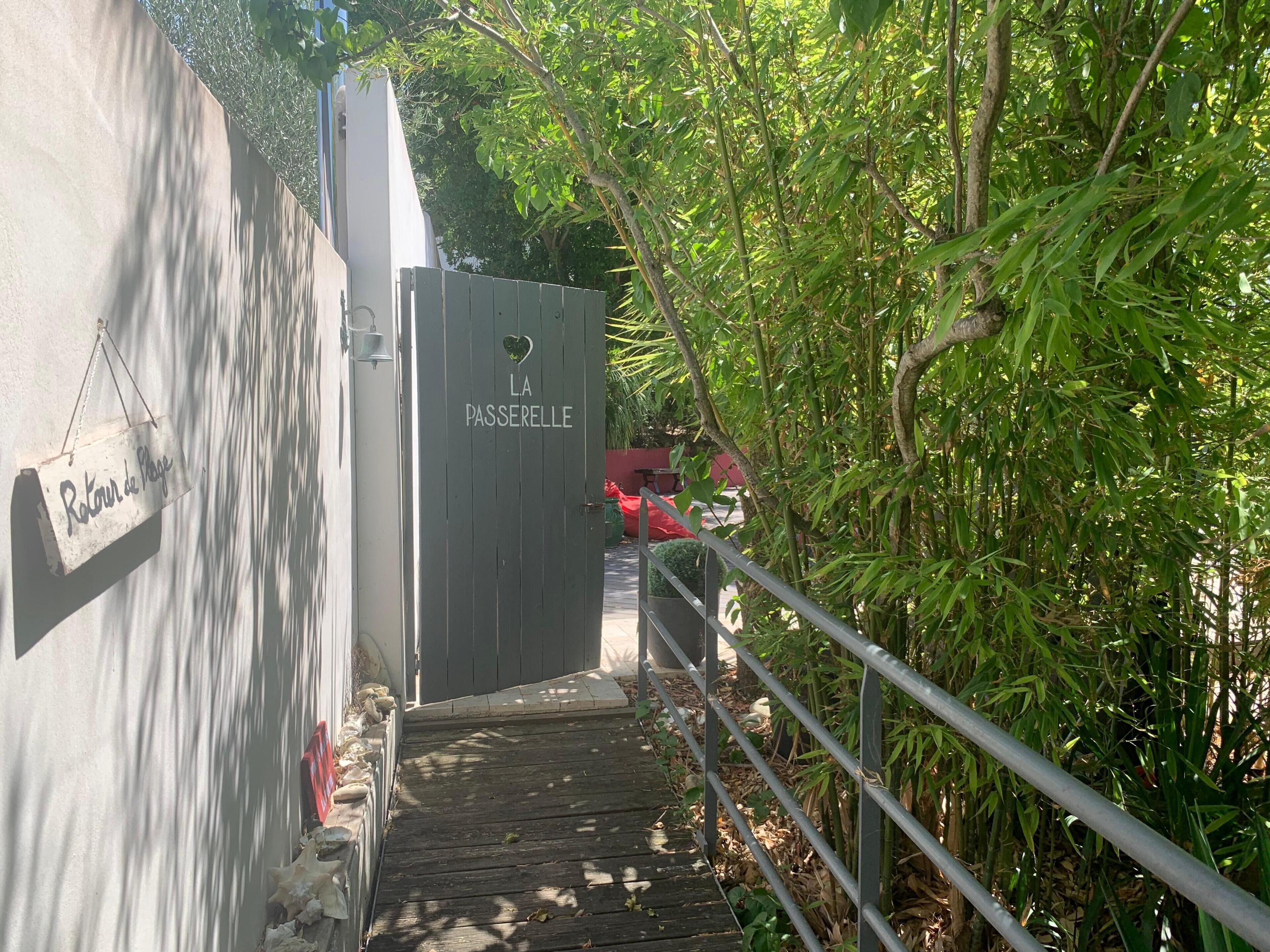Un escalier en bois mène à une porte marquée « LA PASSERELLE », avec une boîte aux lettres et une pancarte sur le mur indiquant « Retour de plage ».