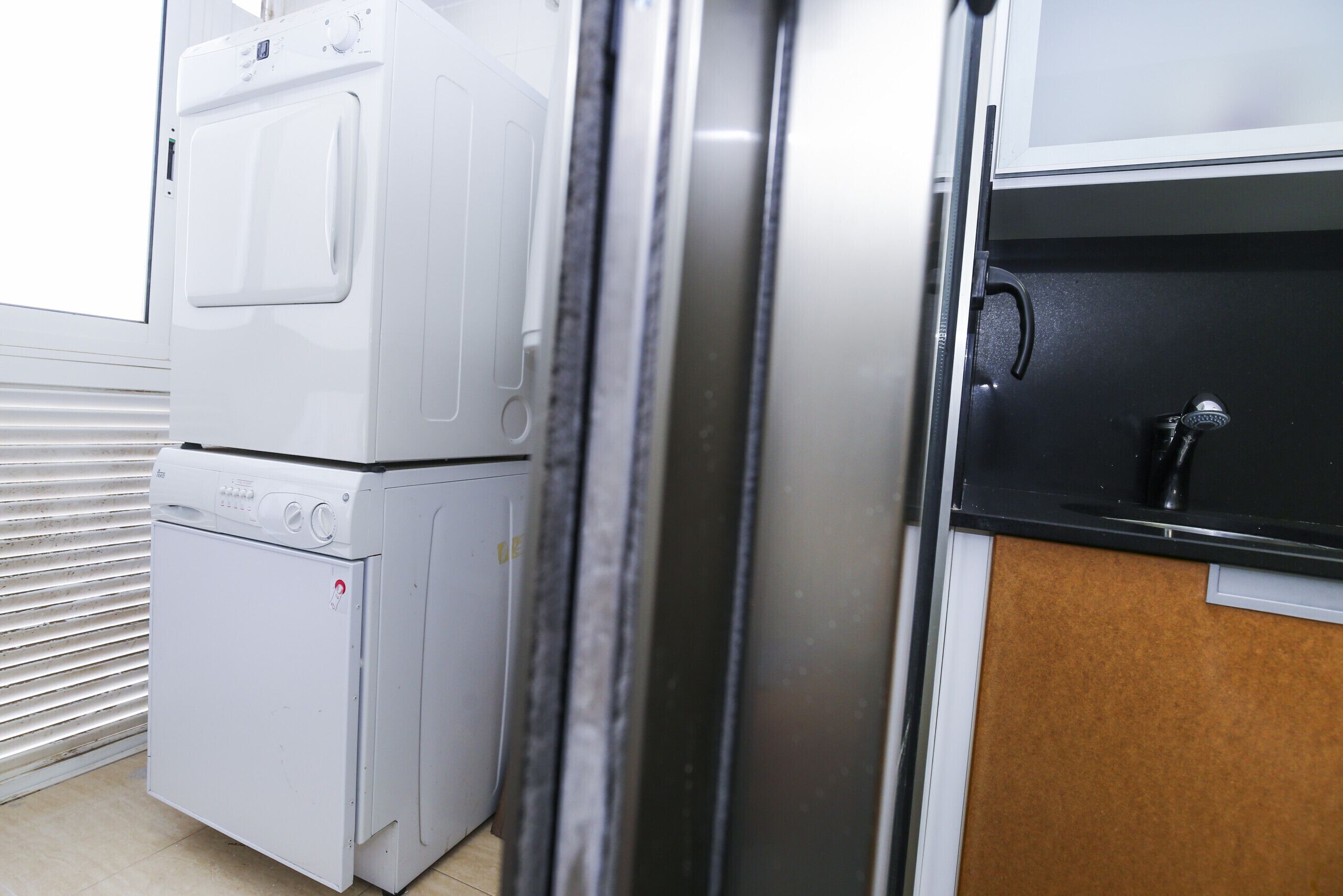Un espace buanderie comprenant une machine à laver et un sèche-linge superposés, un plan de travail noir avec un robinet et une fenêtre avec des stores.
