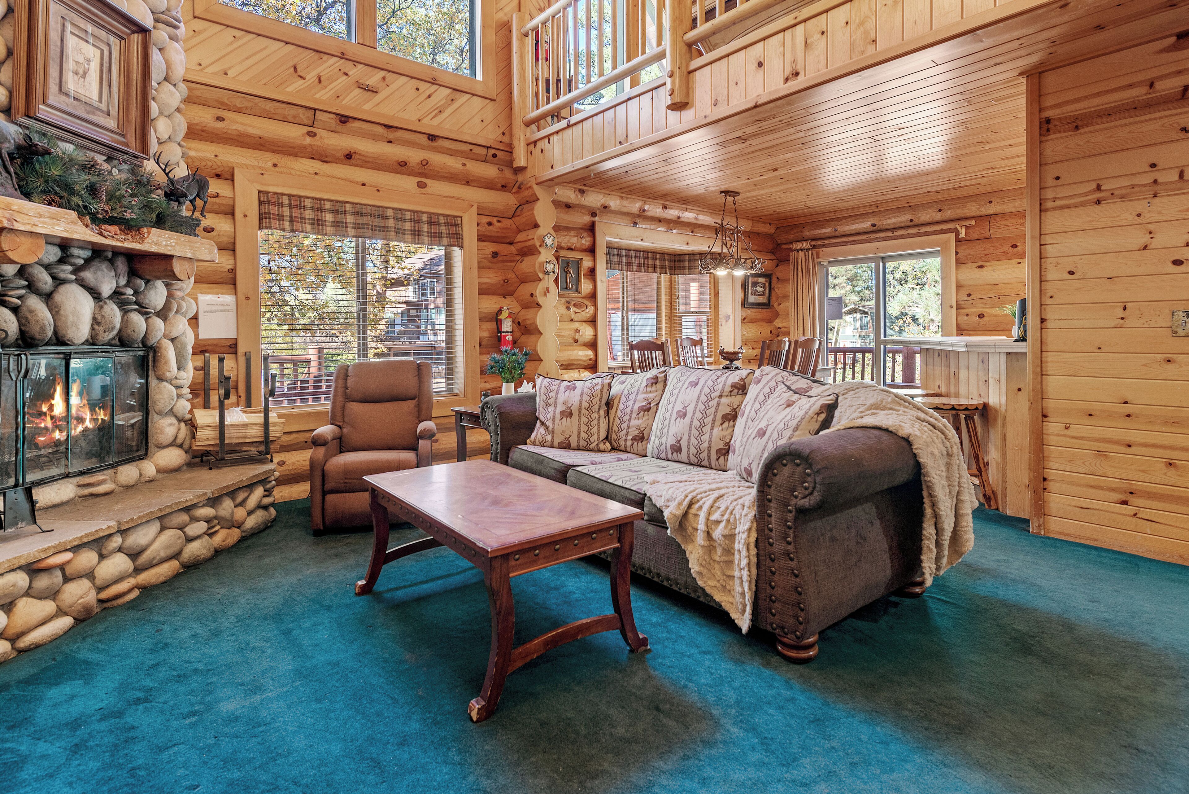 A log cabin interior with a stone fireplace, a sofa, a dining table, and a view of the outside.