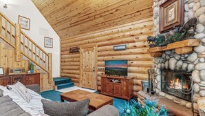 A log cabin interior with a wooden staircase, a fireplace, a television, and a sofa.