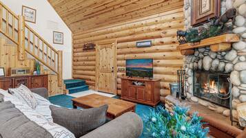 Interior de una cabaña de troncos con escalera de madera, chimenea, televisor y sofá.
