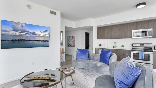 A modern living room with a grey sofa, blue cushions, a glass coffee table, and a large flat-screen TV displaying a scenic view.