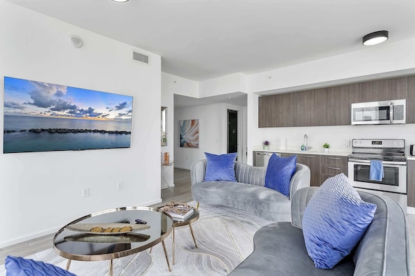 A modern living room with a grey sofa, blue cushions, a glass coffee table, and a large flat-screen TV displaying a scenic view.