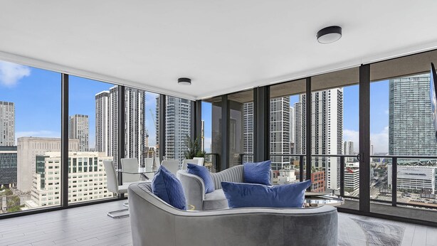 Ein modernes Wohnzimmer mit einem grauen Sofa, blauen Kissen und Blick auf die Stadt durch große Fenster.