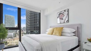 A modern bedroom with a large bed, a city view, and a framed artwork on the wall.