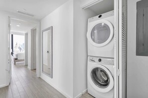 Een wasserette met een wasmachine en droger, een grote spiegel en uitzicht op een stadsgezicht door een geopende deur.