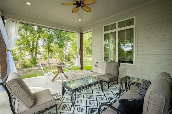 A covered patio with a glass table, cushioned chairs, and a ceiling fan.