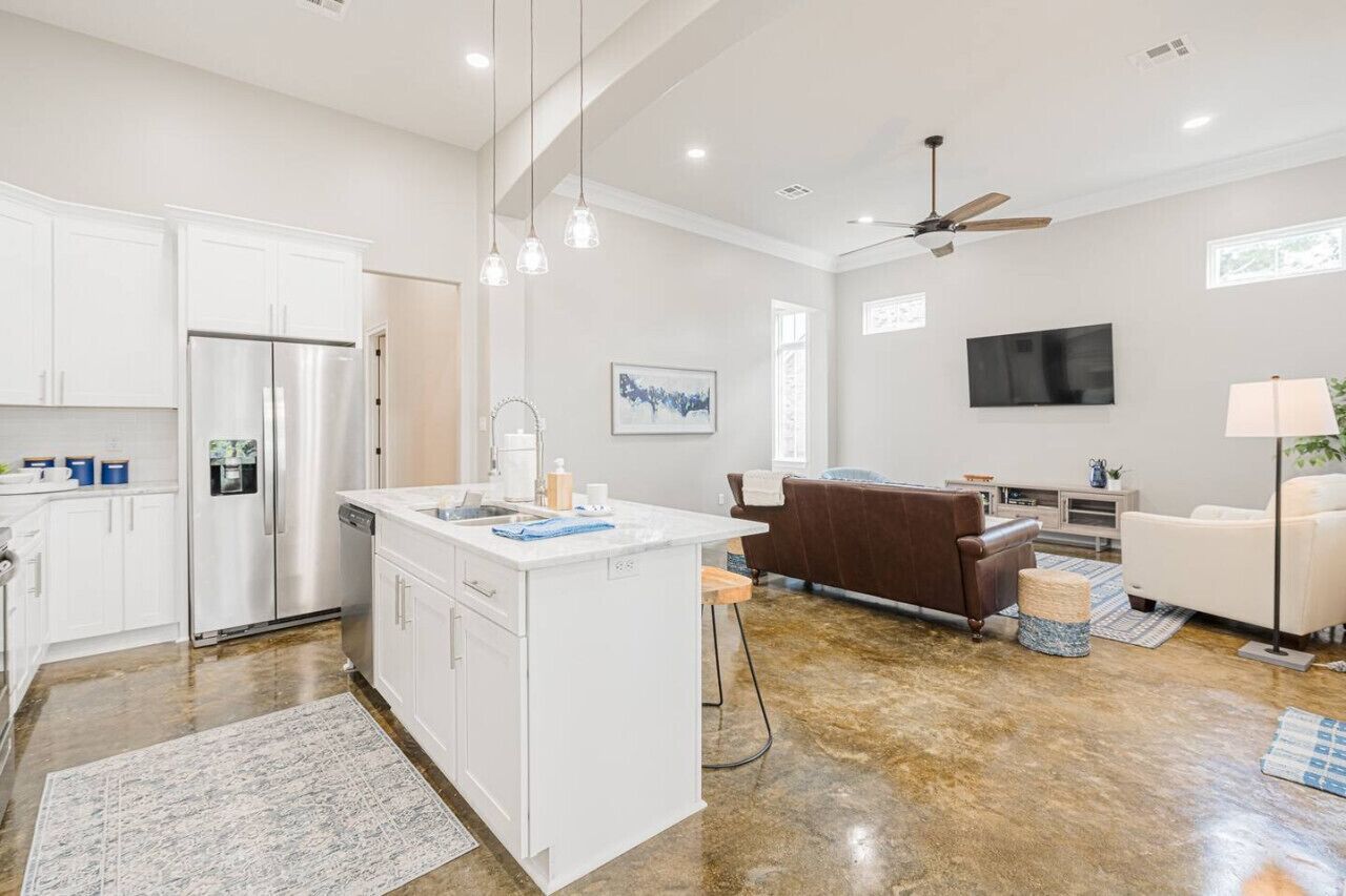 Una cocina moderna con armarios blancos, un refrigerador de acero inoxidable y una isla central. Hay una zona de comedor con un sofá de cuero marrón y un televisor montado en la pared.