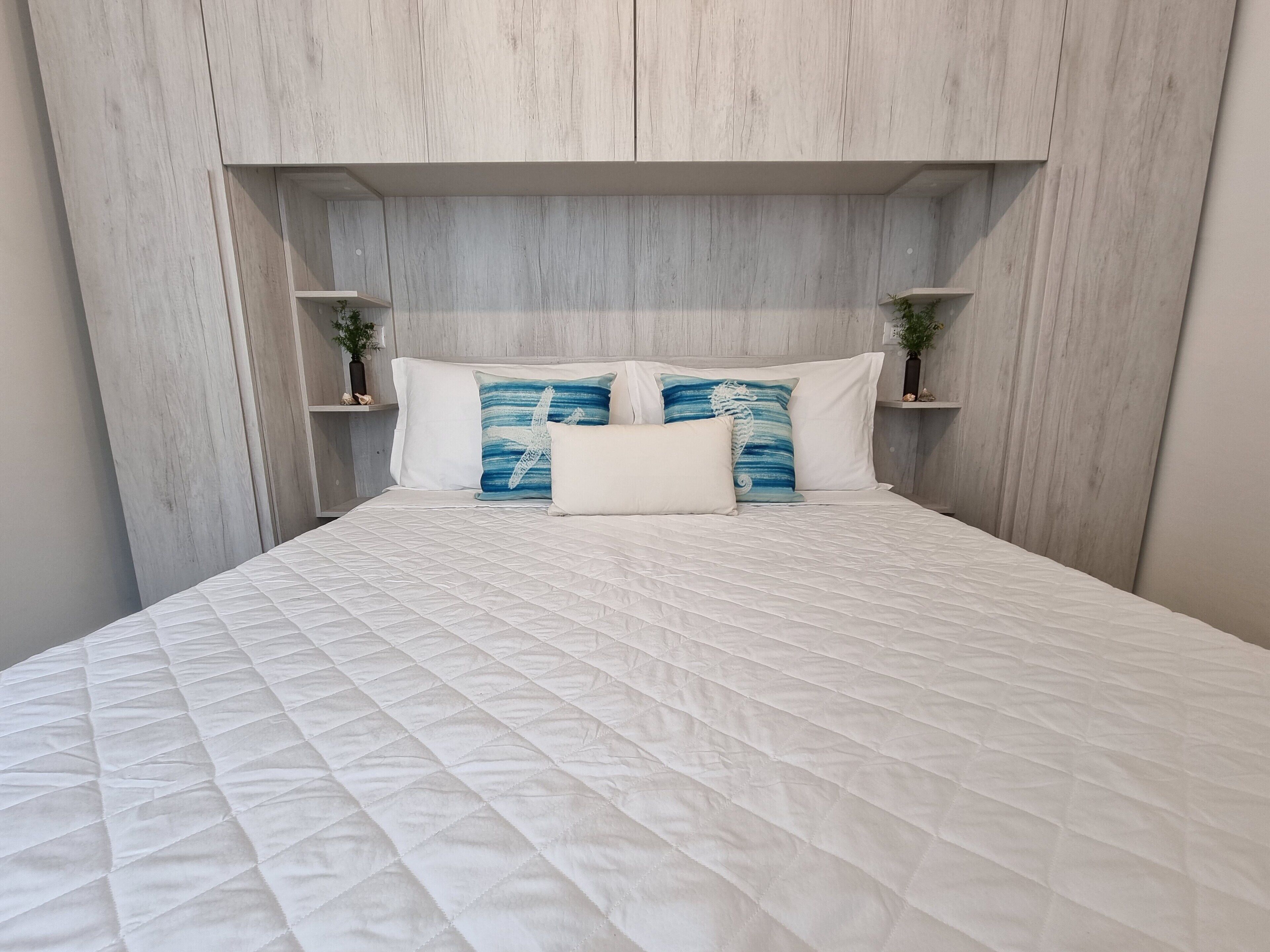 A bed with a quilted white duvet, two pillows with a blue seahorse design, and a headboard with a light wood finish.