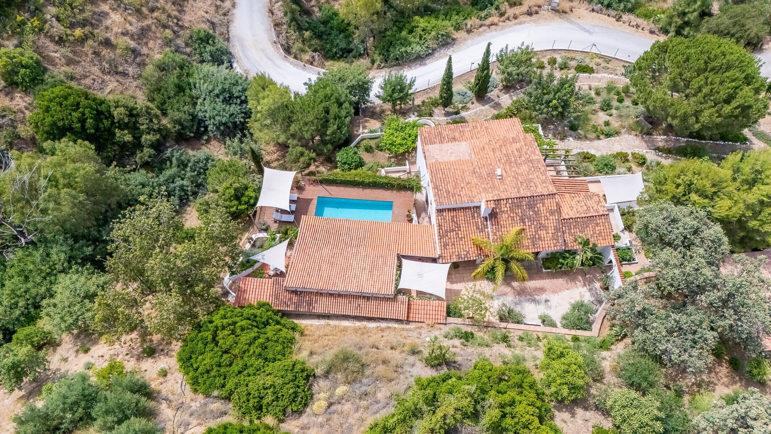 An aerial view of a residential property with a swimming pool, terracotta roofed buildings, and surrounding greenery.