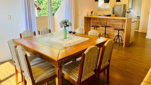 A dining area with a wooden table, chairs, a vase with flowers, and a kitchen in the background. - COBERTURA 04 Suites - RUA COBERTA  (Gramado)
