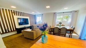 A living room with a sofa, a wooden dining table, a TV, and a vase with flowers. - COBERTURA 04 Suites - RUA COBERTA  (Gramado)