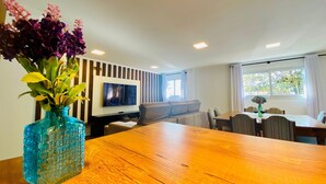 A modern living room with a wooden dining table, a TV mounted on the wall, a sofa, and a window with curtains. - COBERTURA 04 Suites - RUA COBERTA  (Gramado)