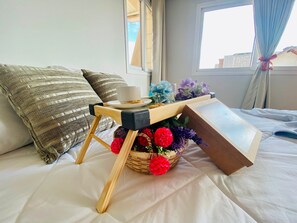 Uma cama com uma mesinha de madeira, uma chávena, um prato com comida e um cesto de flores.