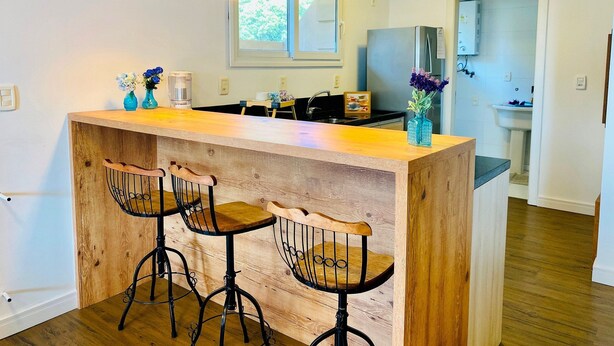 A kitchen with a wooden island, bar stools, pendant lights, and a refrigerator. - COBERTURA 04 Suites - RUA COBERTA  (Gramado)