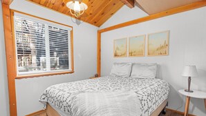 A bedroom with a bed, a window with blinds, a lamp, and two framed pictures on the wall. - Dark Pine Retreat (Big Bear City)