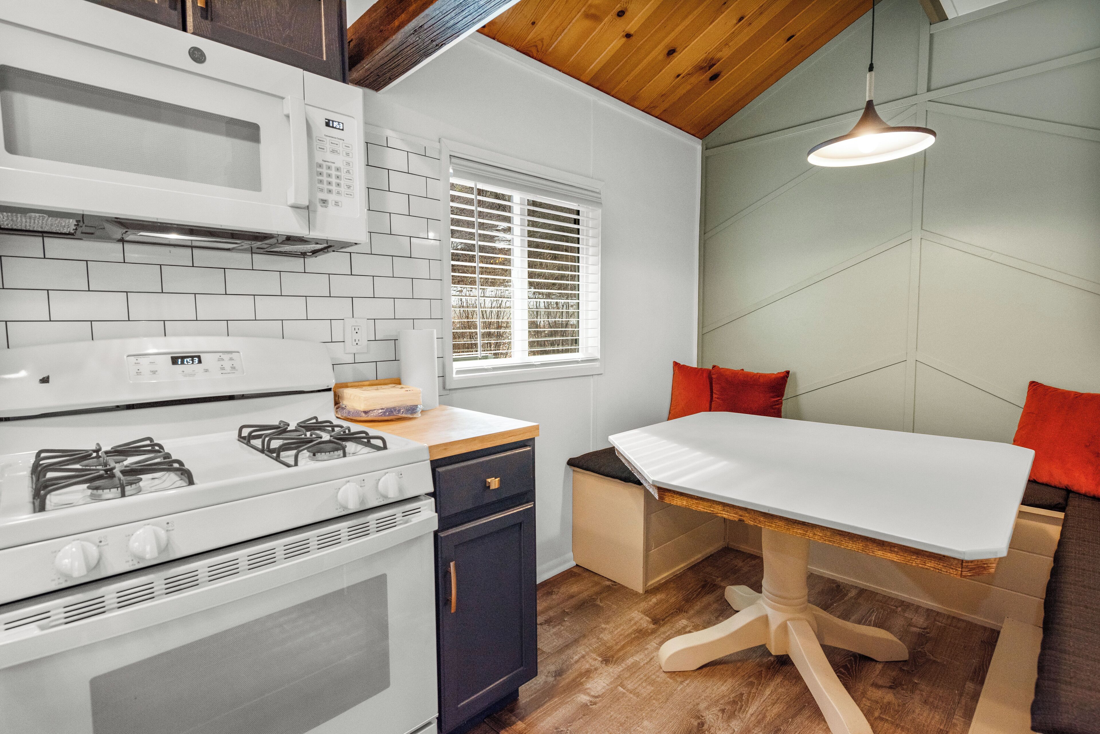 A compact kitchen with a gas stove, microwave, and a small refrigerator. There is a window with blinds, a table with a white tablecloth, and a hanging pendant light.