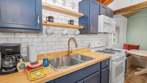 A kitchen with blue cabinets, a white microwave, a gas stove, and wooden shelves above the sink. - Dark Pine Retreat (Big Bear City)