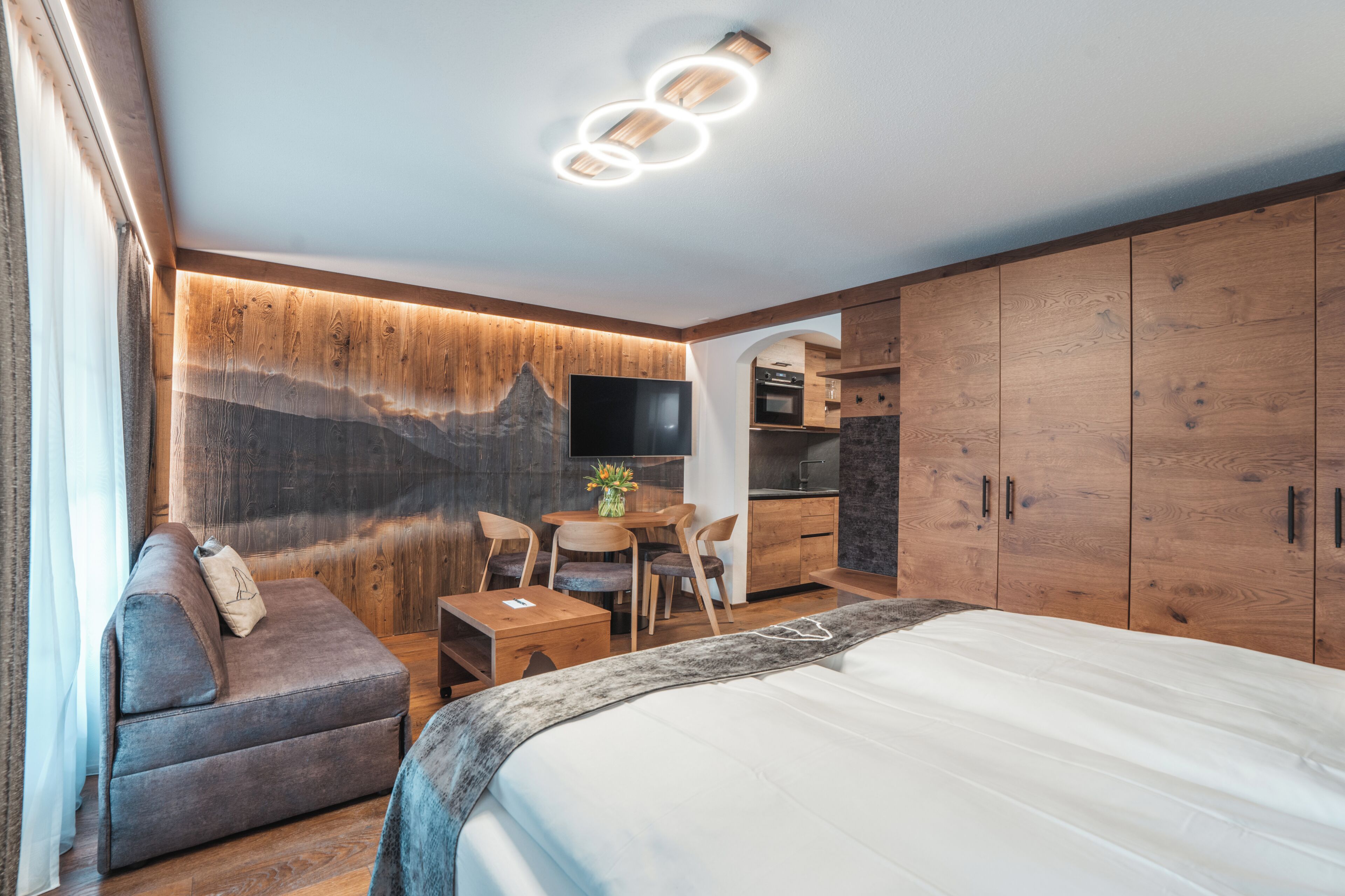 A modern hotel room with a bed, a sofa, a dining table with chairs, a TV, and wooden cabinets.