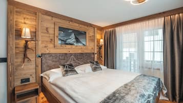 Une chambre à coucher avec une tête de lit en bois, un lit avec une literie blanche, deux lampes de chevet, un tableau encadré représentant une montagne et une fenêtre avec des rideaux.